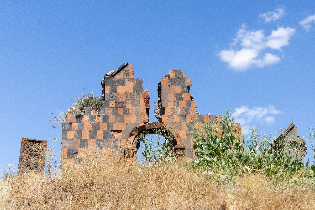 The checkerboard tuff-and-basalt facade of Havuts Tar standing above dry summer grass, with a khachkar at its left.