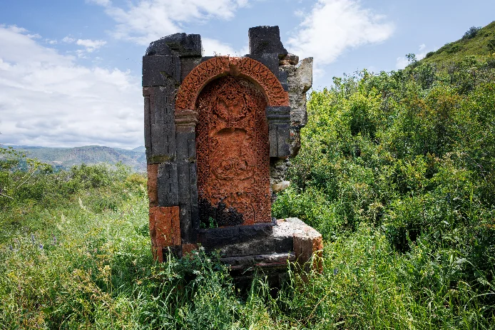 A khachkar carved in orange tuff, set in a dark stone frame and surrounded by tall summer grass.